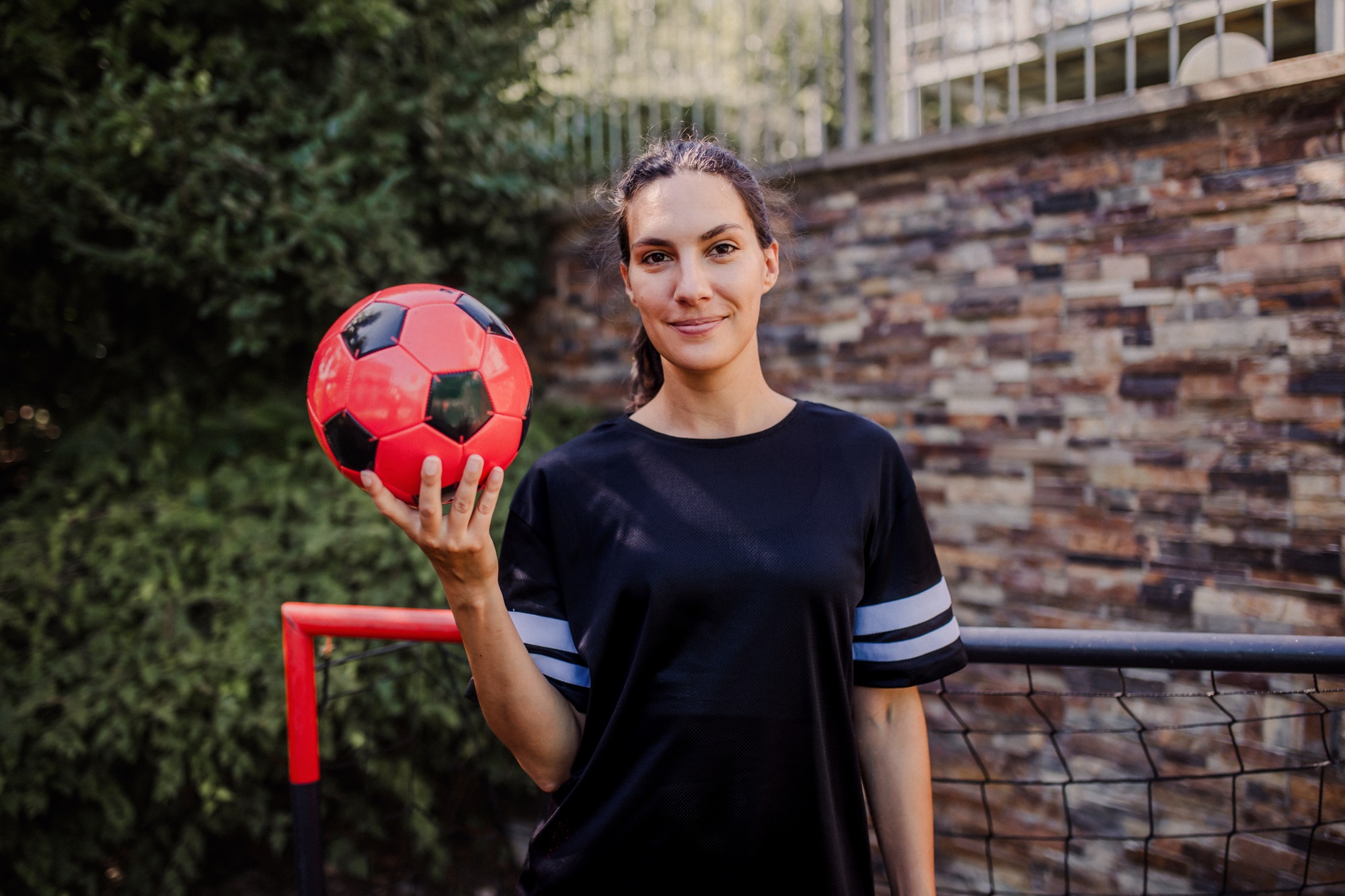 Beautiful female football player standing by football goal with ball in her hand. The woman plays
